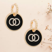 Load image into Gallery viewer, Gold-tone twisted hoop earrings with round black epoxy charms and interlocking rhinestone rings on a floral background
